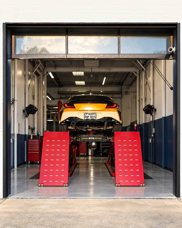 A sports car raised on an alignment lift, seen from behind through an open bay door.