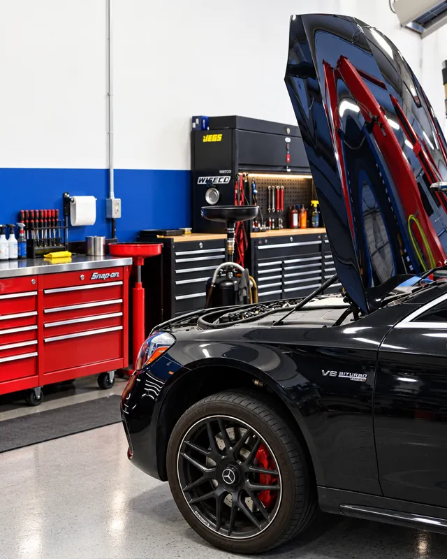 A black sedan with its hood up beside red and black tool chests.