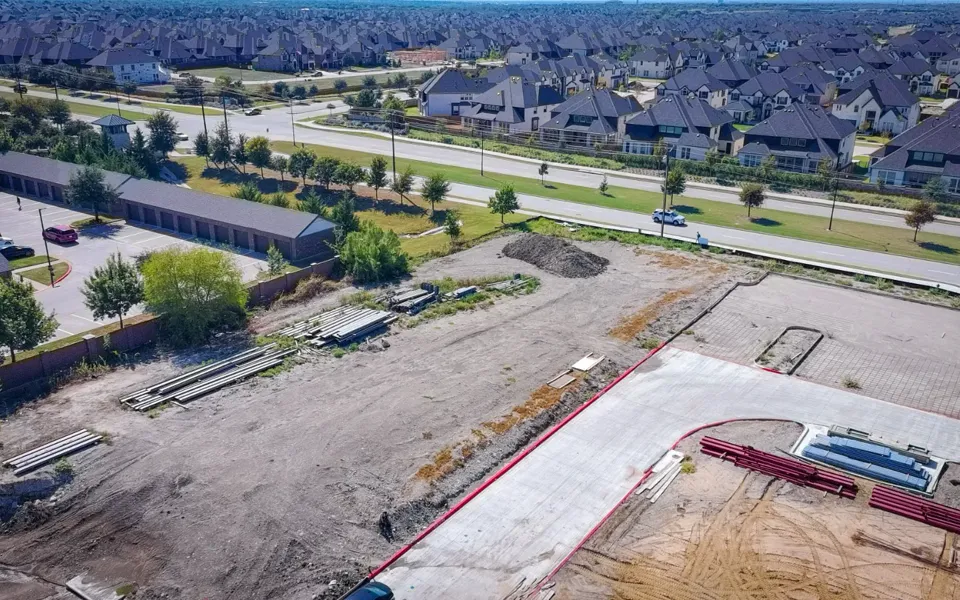 Aerial view of the graded McKinney parcel in October 2026, with stacked building steel, a poured concrete drive and housing beyond.