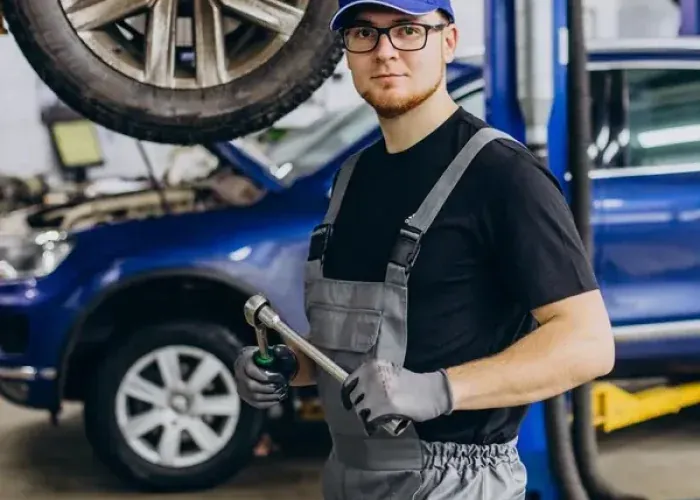 Auto technician in work overalls and gloves holding a wrench, with a car raised on a service lift behind